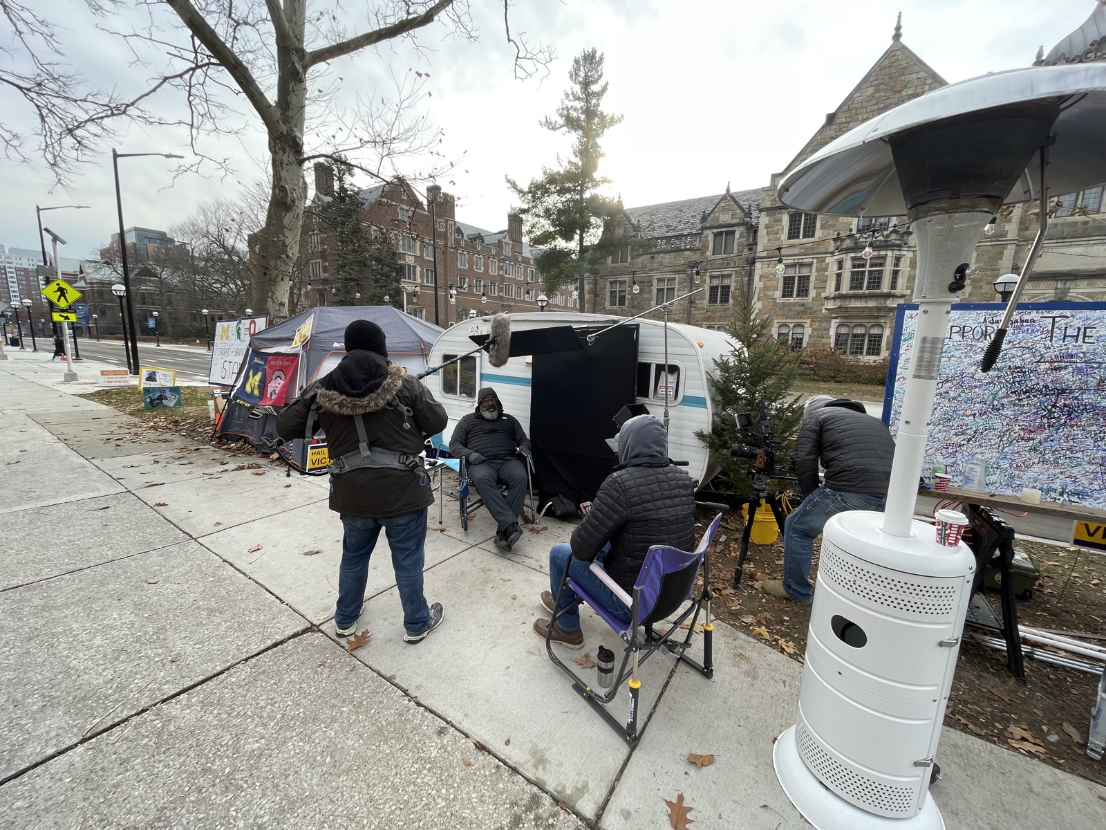 Film crew with boom mic and camera shooting Jon Vaughn outside his tent and camper on the University of Michigan campus during Hail To The Victims