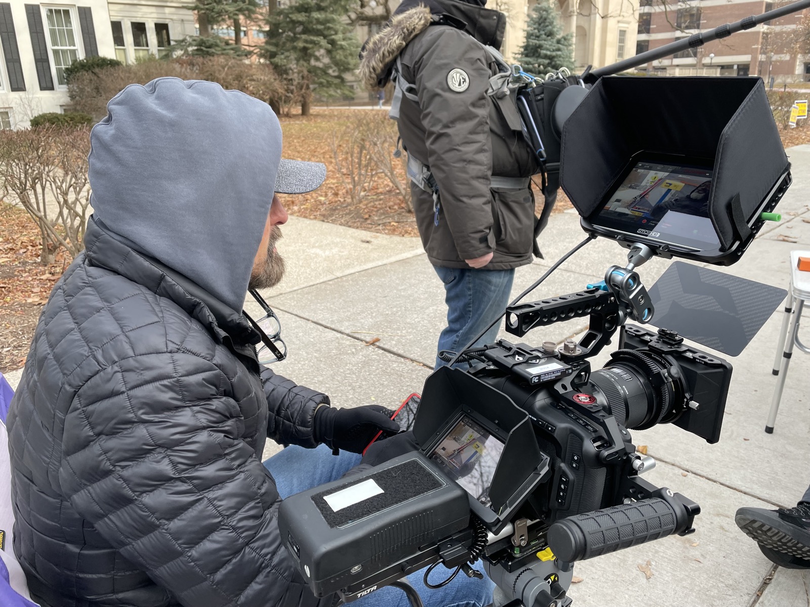 Camera operator kneeling behind a rigged cinema camera with matte box and monitors on campus during the Hail To The Victims shoot